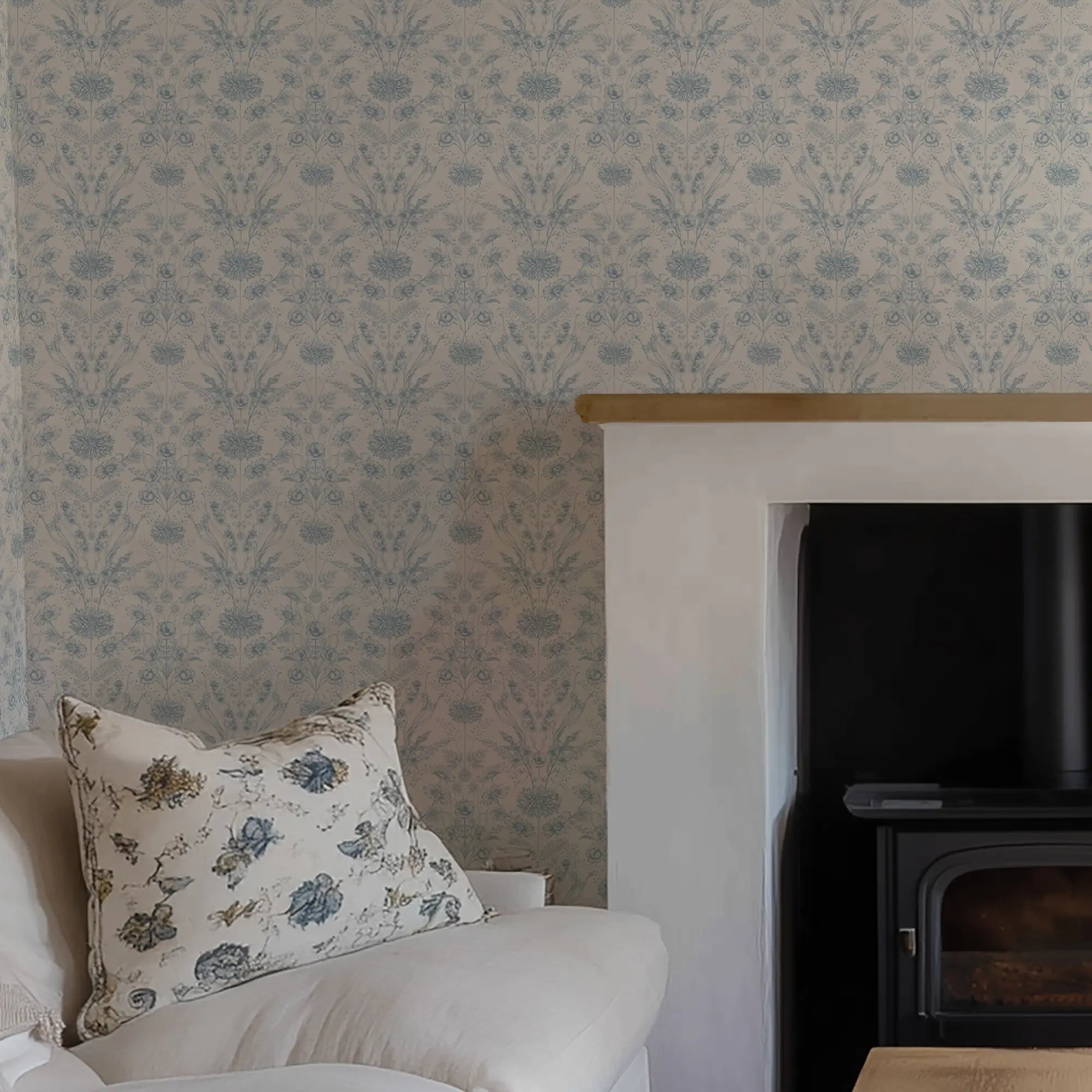Floral pillow on a sofa against a patterned wall with blue flowers next to a fireplace.
