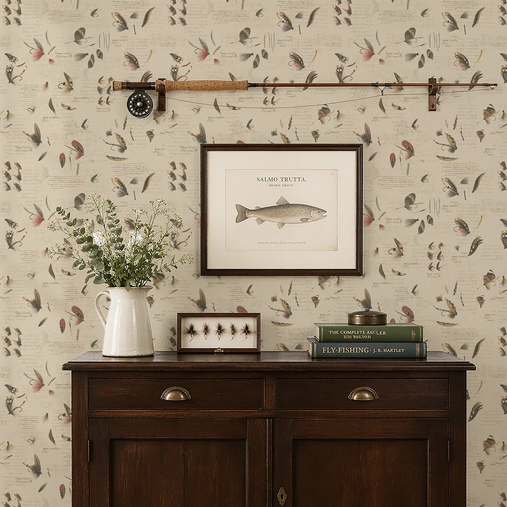 Wall with decorative wallpaper featuring fishing themed design, a wooden sideboard with books and a vase, and framed pictures.