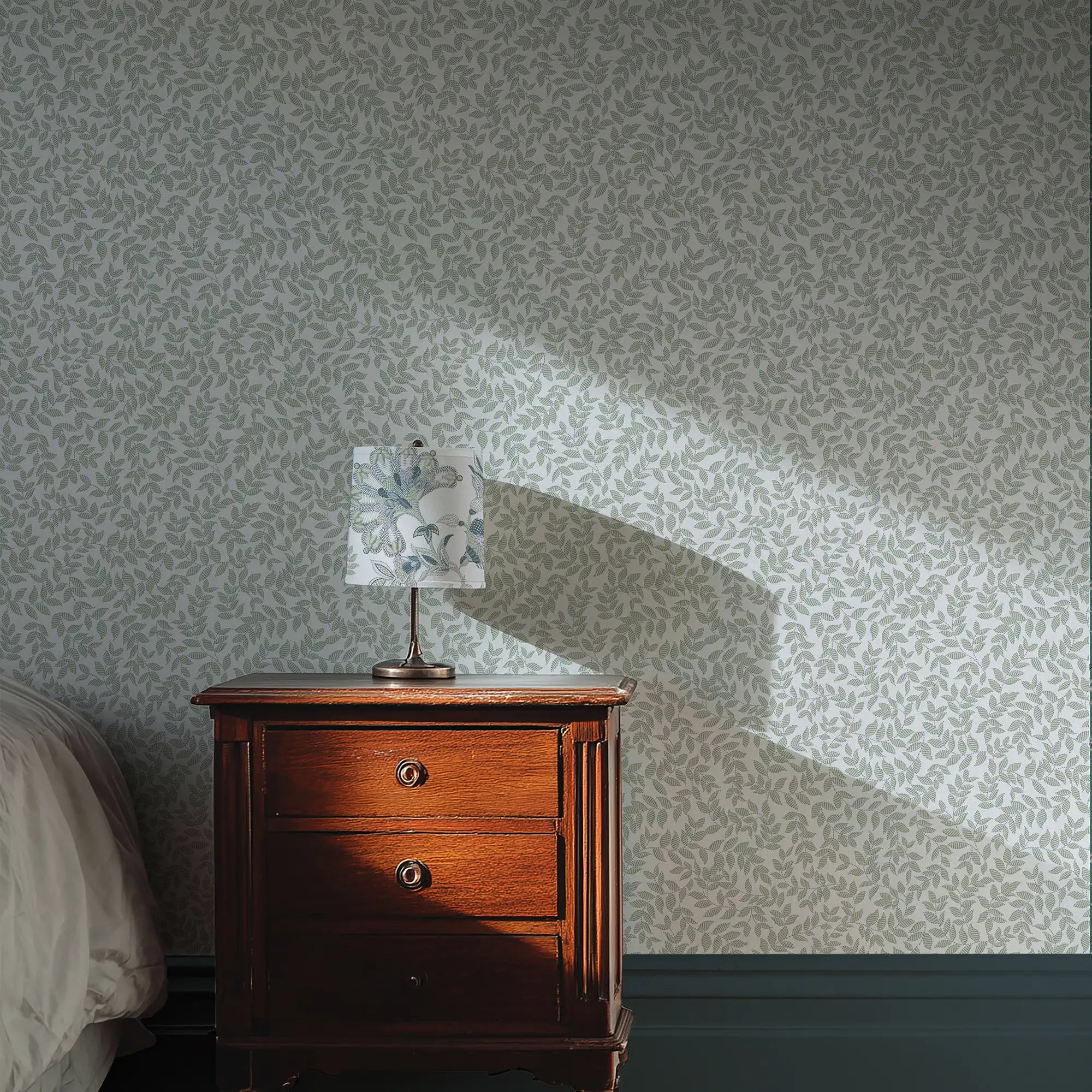 Wooden nightstand with a lamp against a patterned wall with trailing leaves.